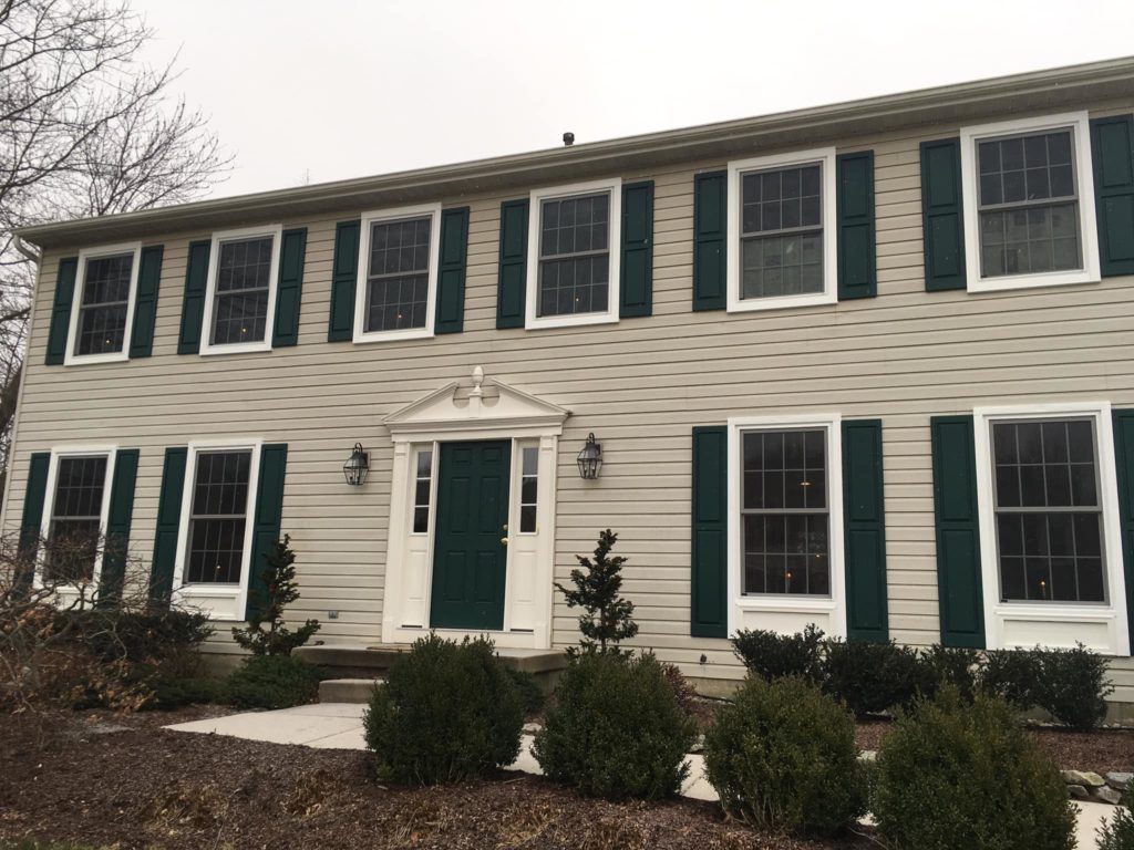 Large colonial home with tan siding and dark green, solid shutters. Multiple white, double hung replacement windows with grids. Windows with candles.